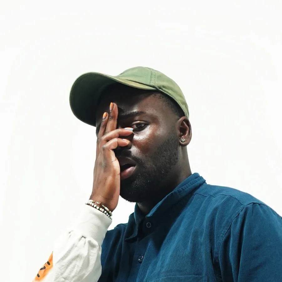 A young man with dark skin wearing a green cap, blue shirt, and earrings, partly covering his face with his hand, standing against a plain white background.