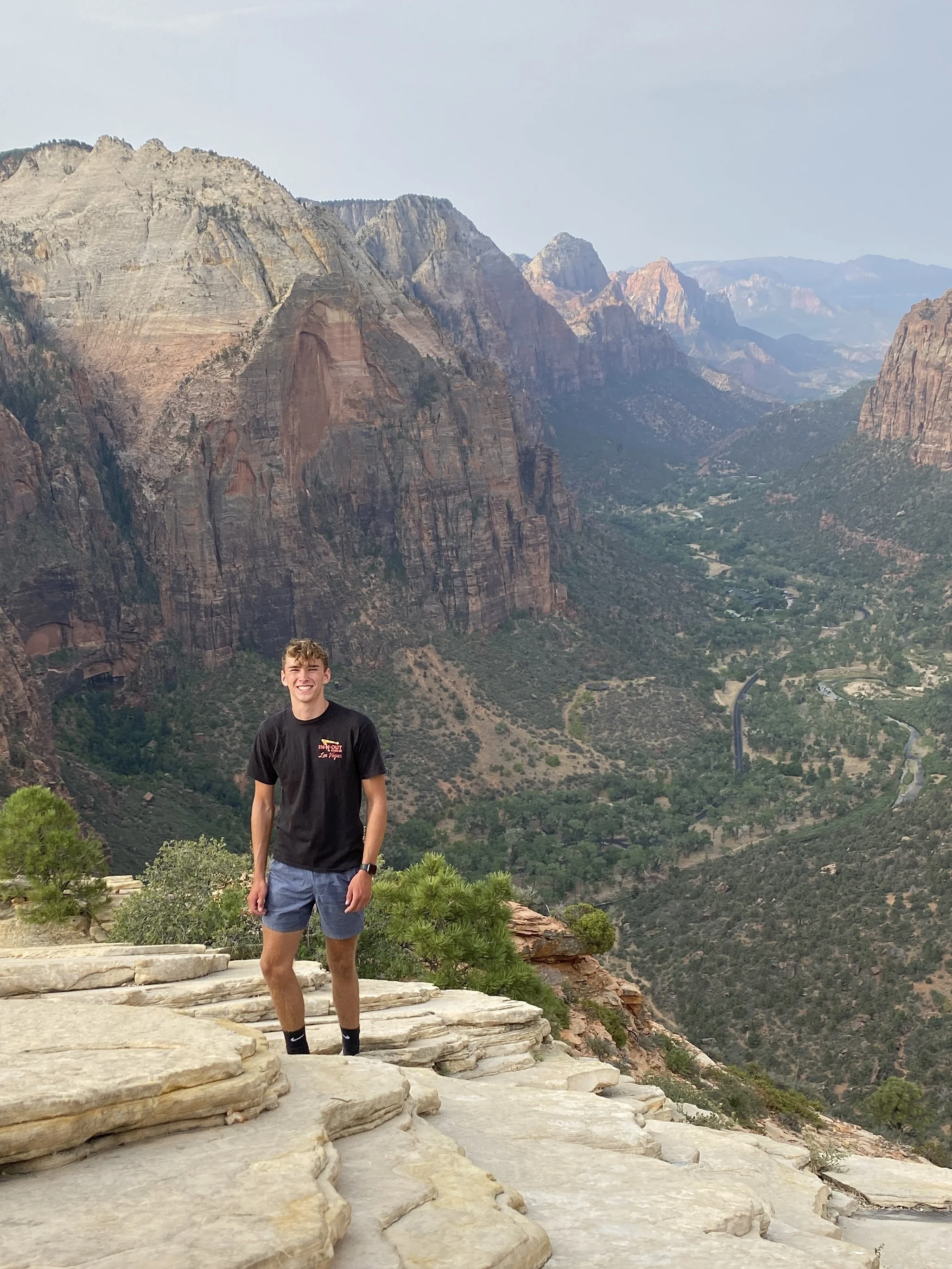 A young man in a black t-shirt and blue shorts standing on a rocky ledge in front of a vast canyon landscape with tall cliffs and a river below.