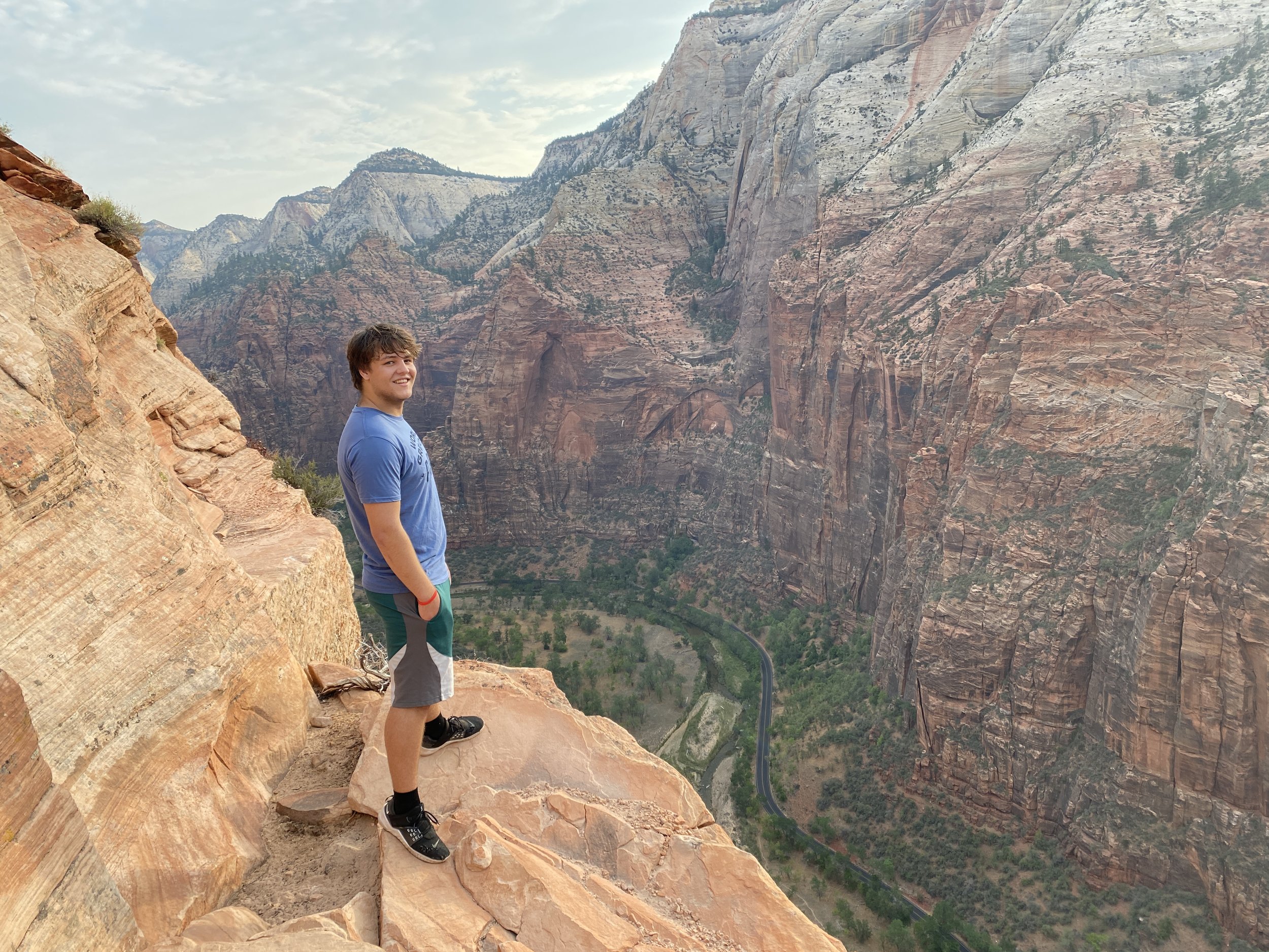 A young man standing on a rocky ledge overlooking Zion Canyon with steep red cliffs and a winding road below.