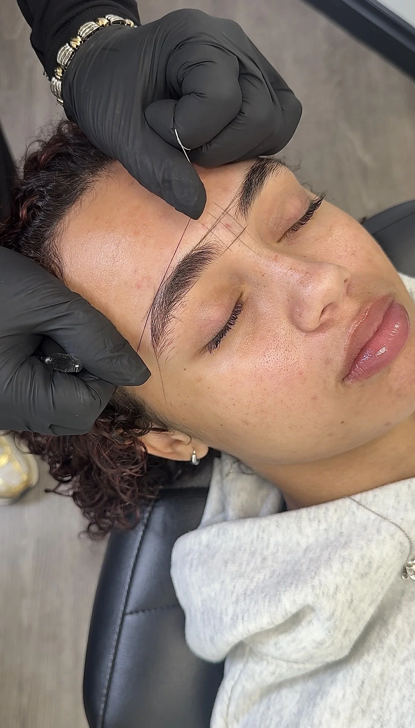 A woman with dark hair and earrings receives a microblading tattoo on her eyebrows from a technician wearing black gloves.