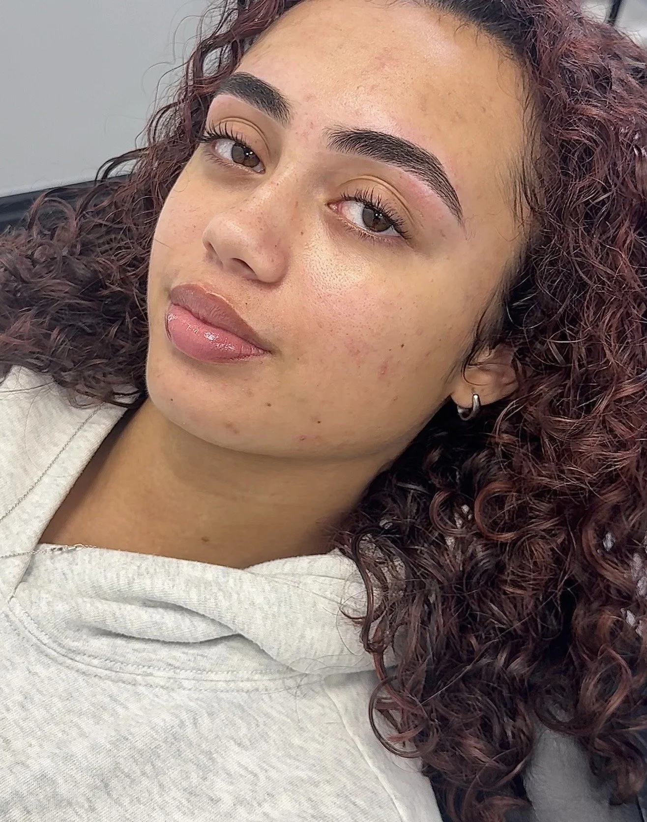 Close-up photo of a woman with curly burgundy hair, with natural makeup, light brown eyes, and well groomed eyebrows looking at the camera.