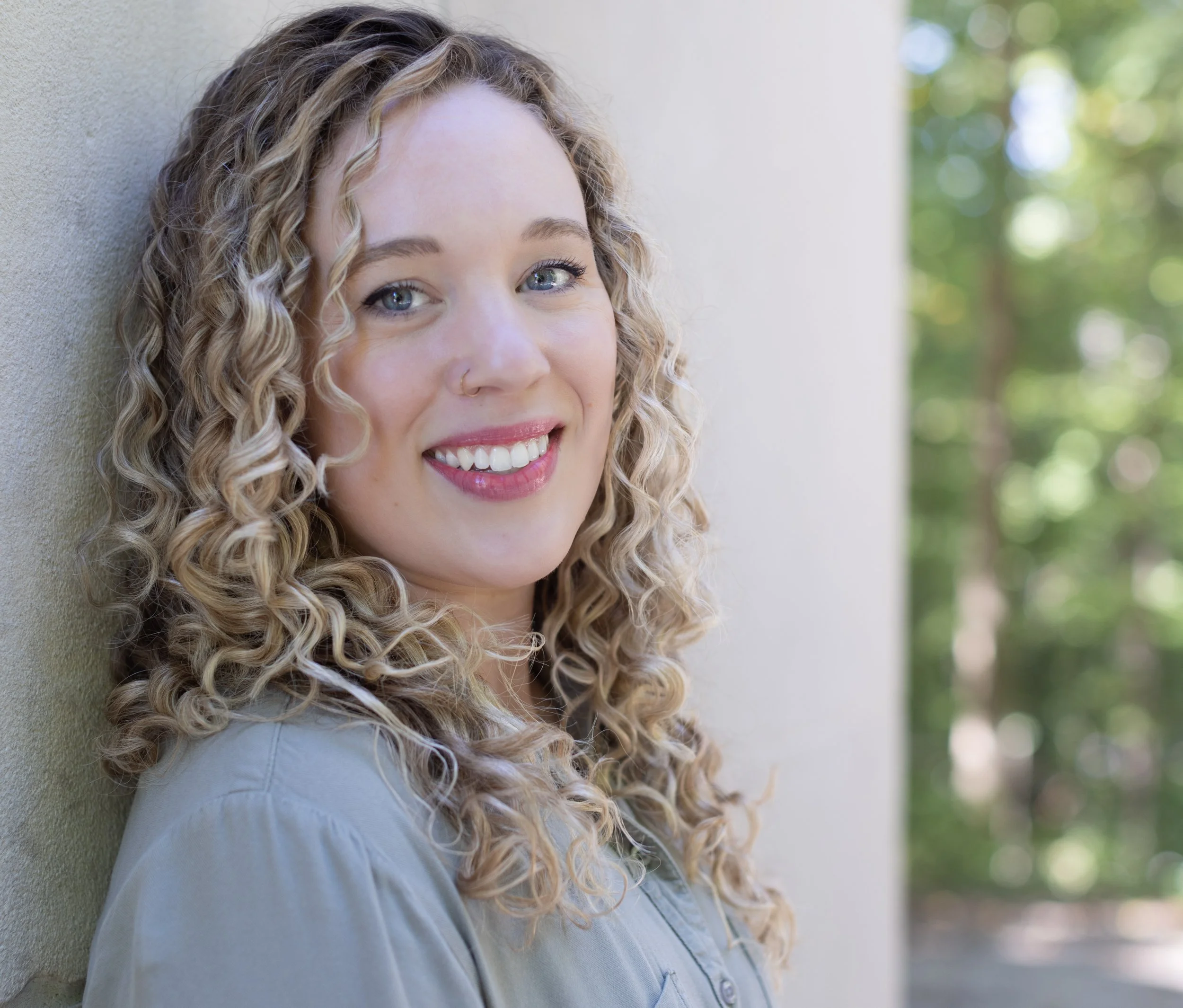 A young woman with curly blonde hair and blue eyes smiling, leaning against a beige wall with green trees in the background.