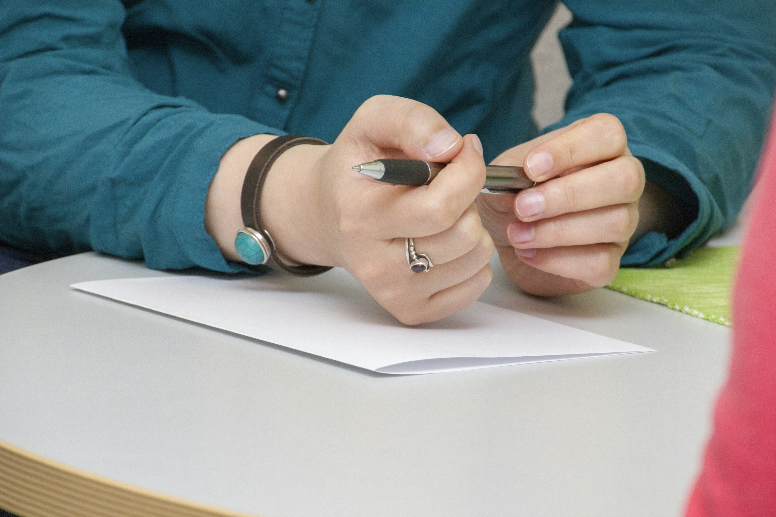 Close-up of someone writing with a pen on a white sheet of paper on a table. The person wears a ring and a bracelet with a turquoise stone.