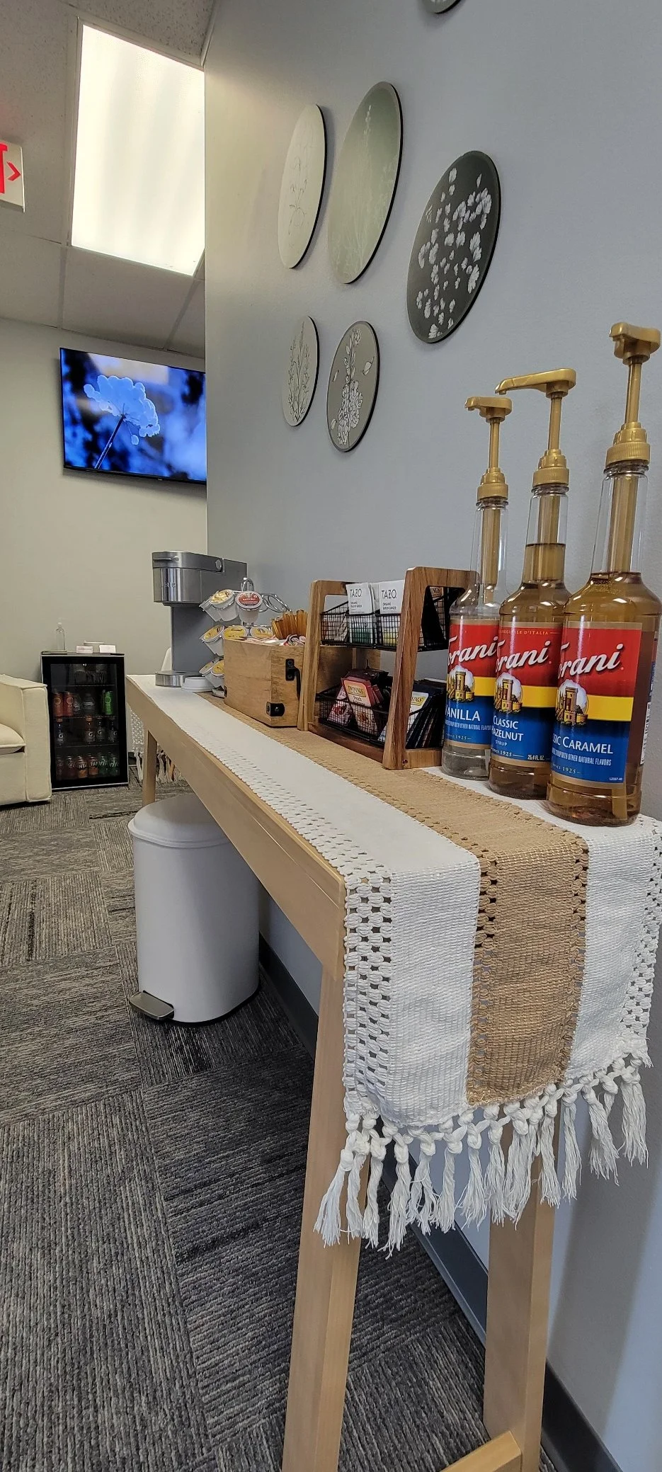A coffee station with four flavored syrup bottles, a wooden caddy holding tea bags, a coffee machine, and a small refrigerator in the background. The station is covered with a decorative table runner and is located in a room with gray carpet and white walls, decorated with round wall art.