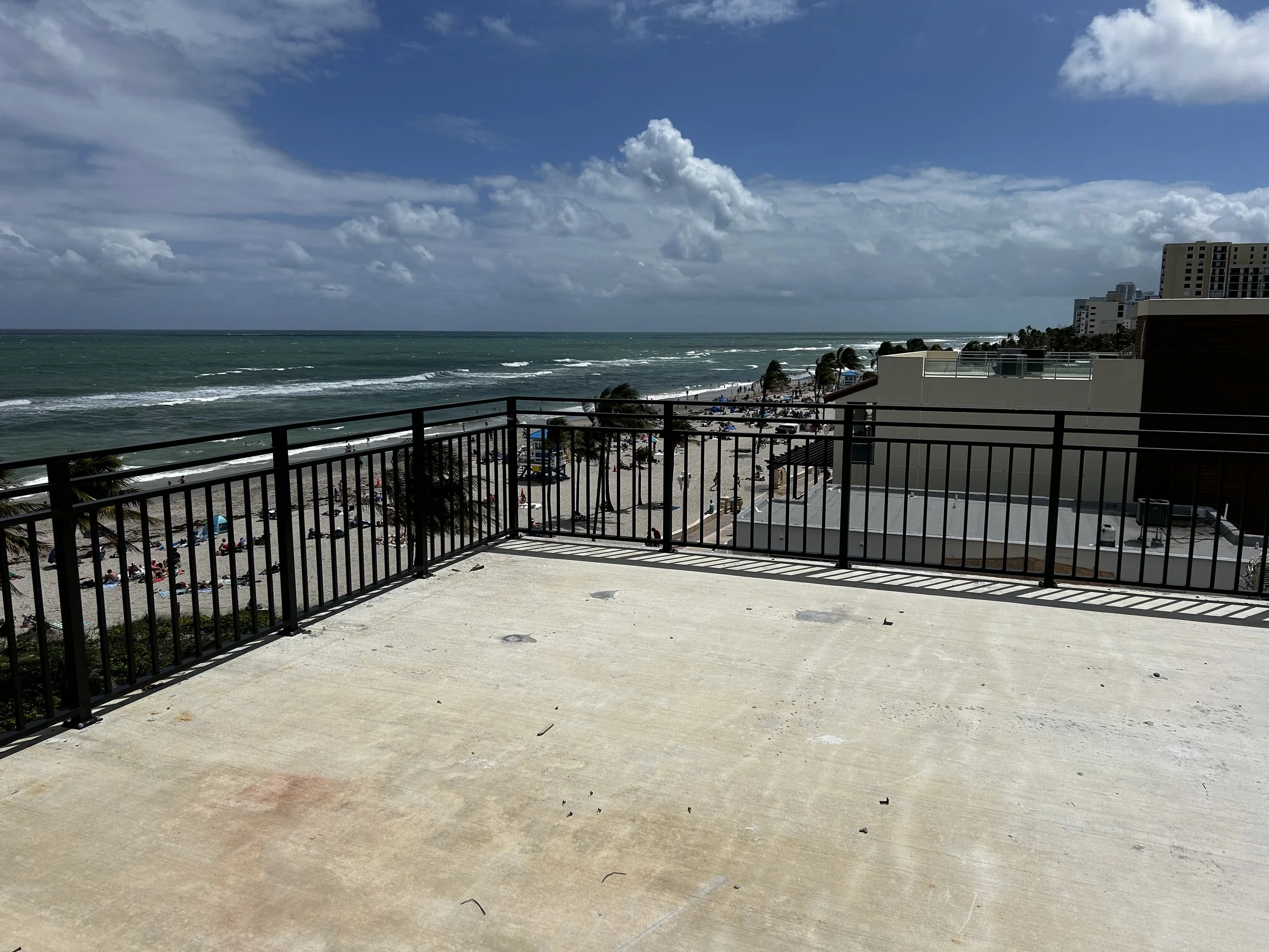 Vista desde un balcón con barandilla de metal, mostrando playa con arena, personas, palmeras, edificios y el mar con olas bajo un cielo parcialmente nublado.