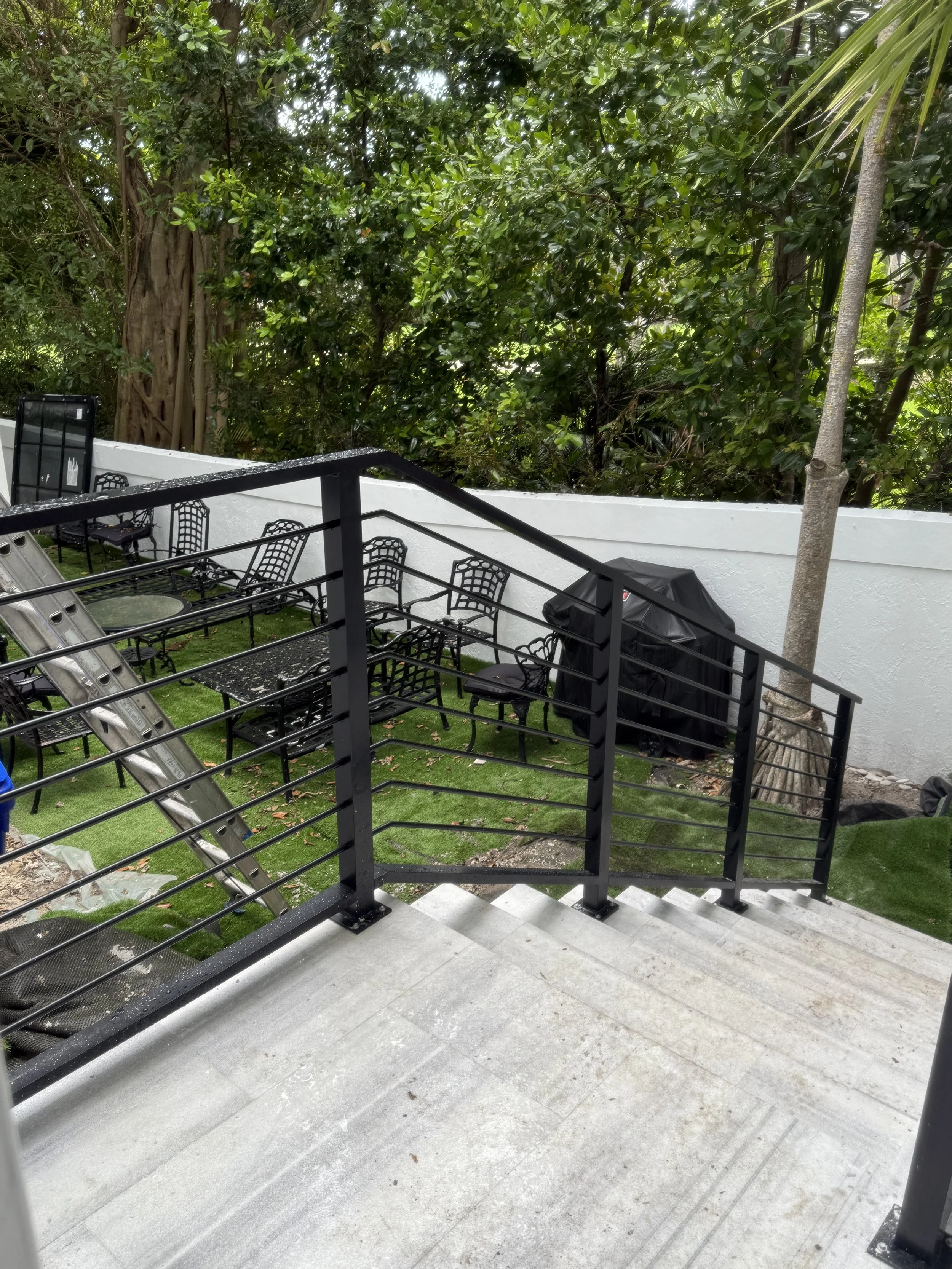 Escalera de concreto con barandal de metal negro, área de descanso con sillas de malla negra y una mesa, espacio para barbacoa cubierto, cercanía a un muro blanco y árboles grandes con follaje verde.