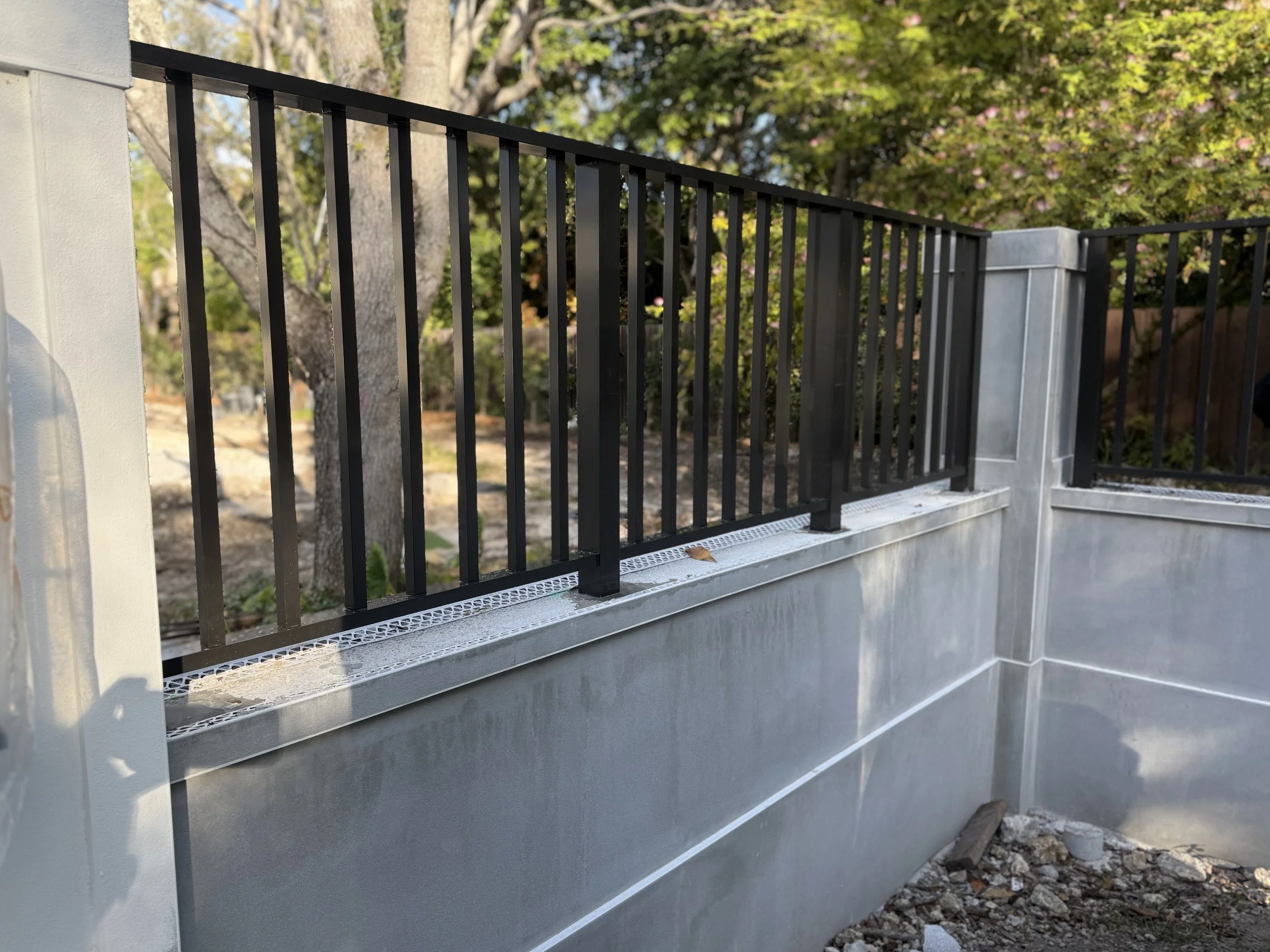 Barandal negro instalado sobre una base de concreto en un área exterior, con árboles y vegetación en el fondo.