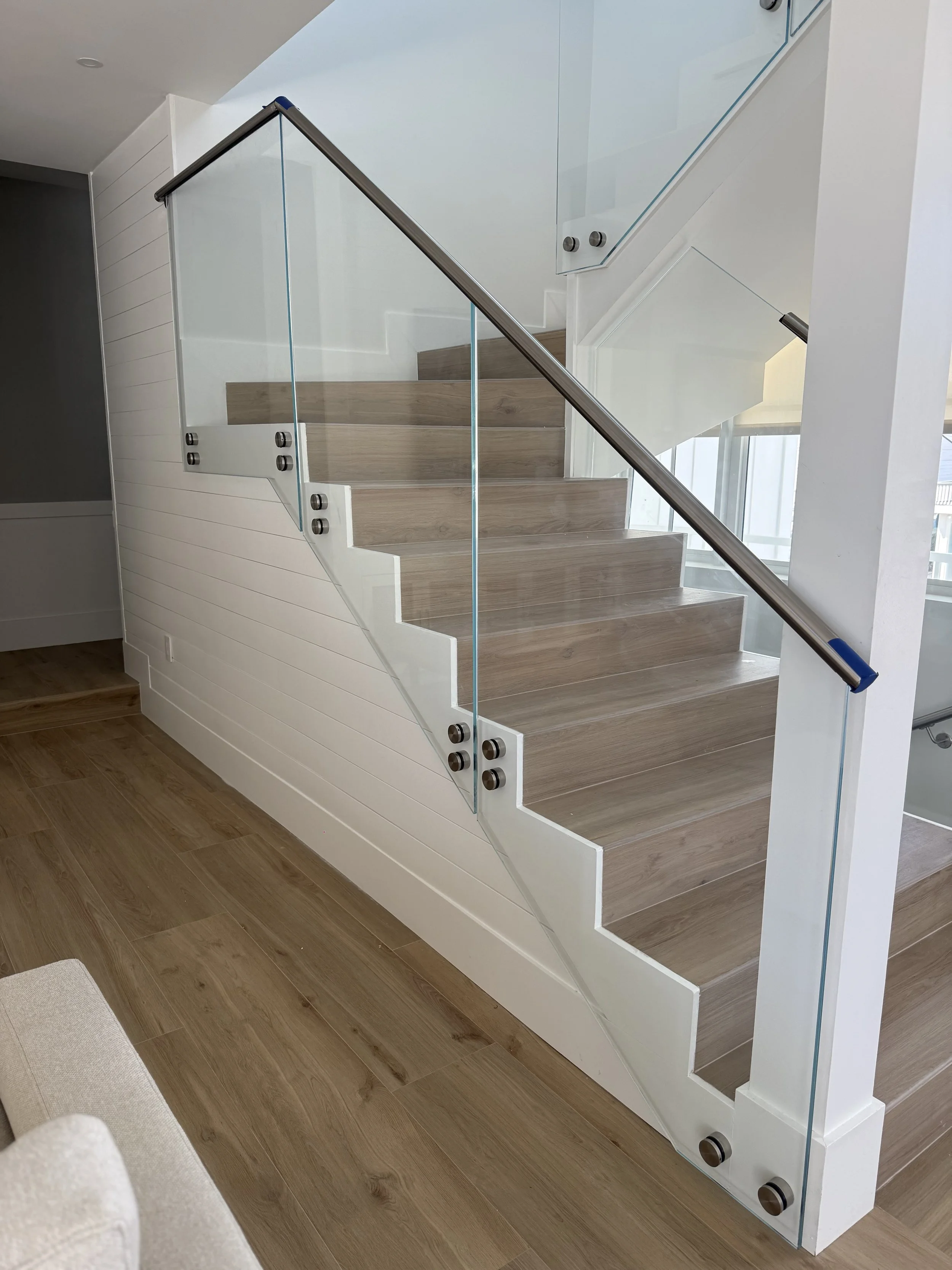 Escalera moderna con barandilla de cristal y pasamanos de acero, en un espacio interior con paredes blancas y suelo de madera.