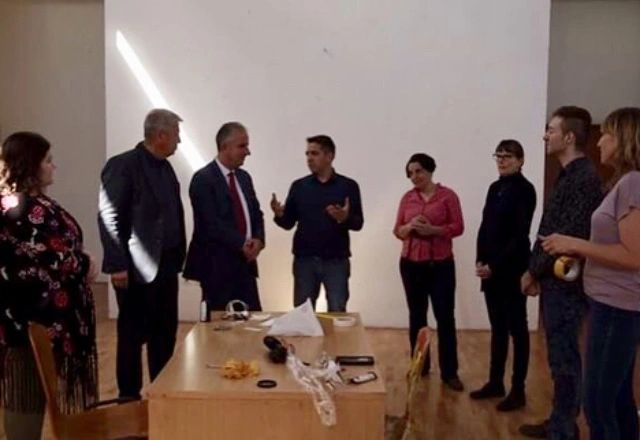 Group of people standing in a room, engaged in conversation, with a table holding various items in front of them.