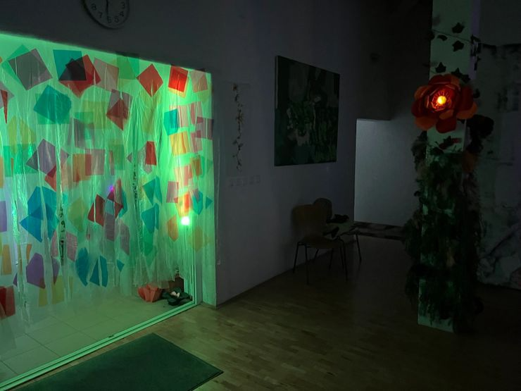 Dark room with a colorful, illuminated curtain with paper shapes, a large artificial flower with lights, a chair, and paintings on the wall.