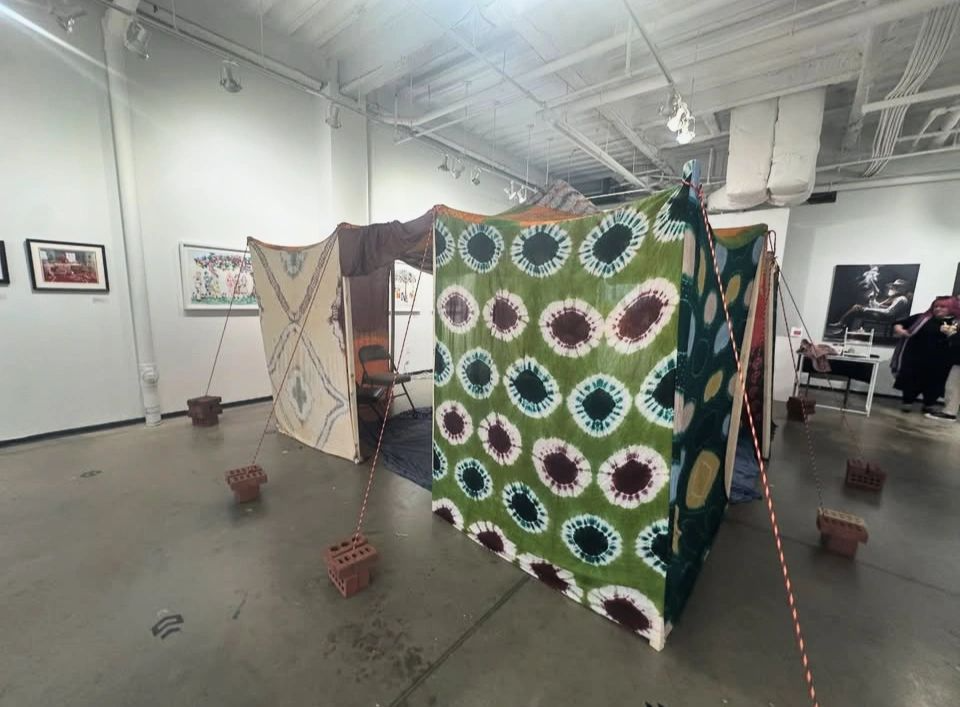 Art installation of four fabric panels with tie-dye patterns, secured with red and white striped ropes anchored by small brick bases in a gallery space with framed artwork on white walls.