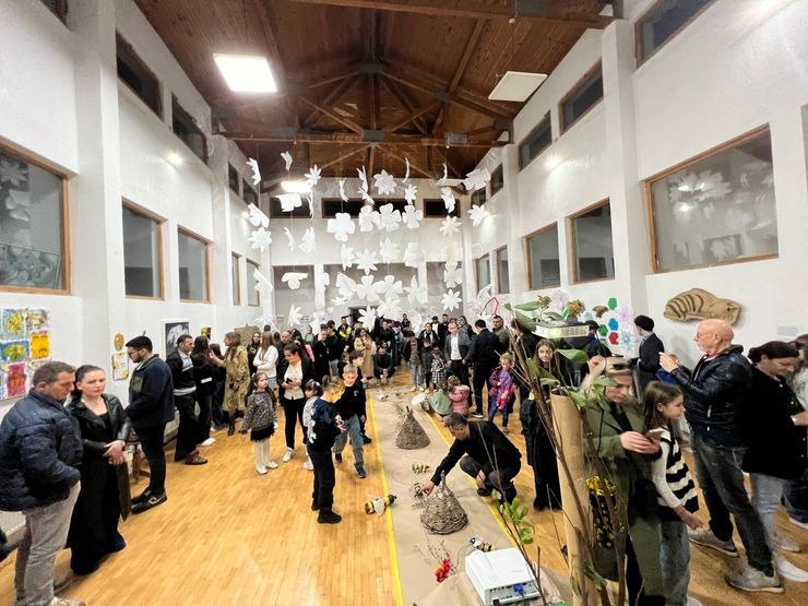 People gather in a large indoor hall with wooden floors and high vaulted ceiling, decorated with hanging paper leaves and other crafts, at an art or craft fair.