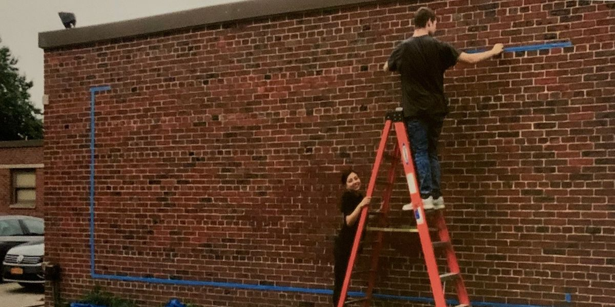 Two people painting blue tape on a brick wall in preparation for a mural or art project, one person standing on a ladder and the other standing on the ground holding the ladder.