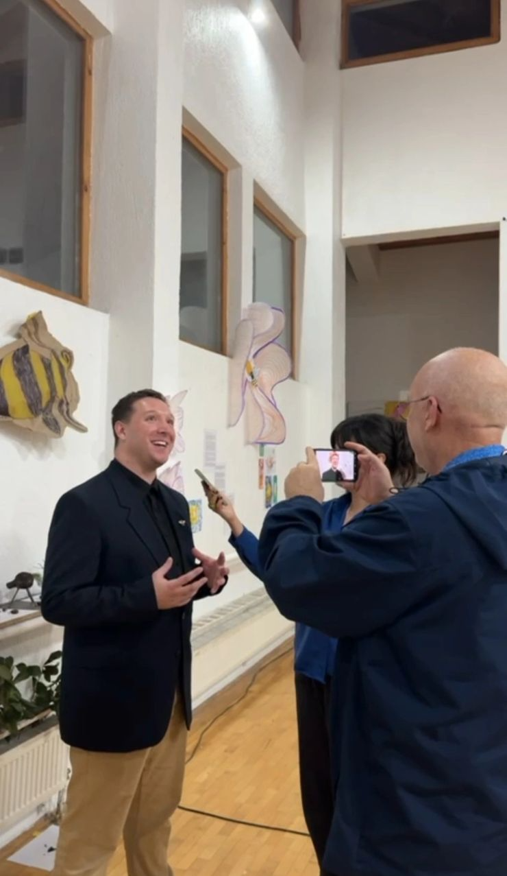 A man in a dark blazer and light-colored pants is speaking or presenting while three people take photos or videos of him in an indoor space with white walls, windows, and artwork on display.