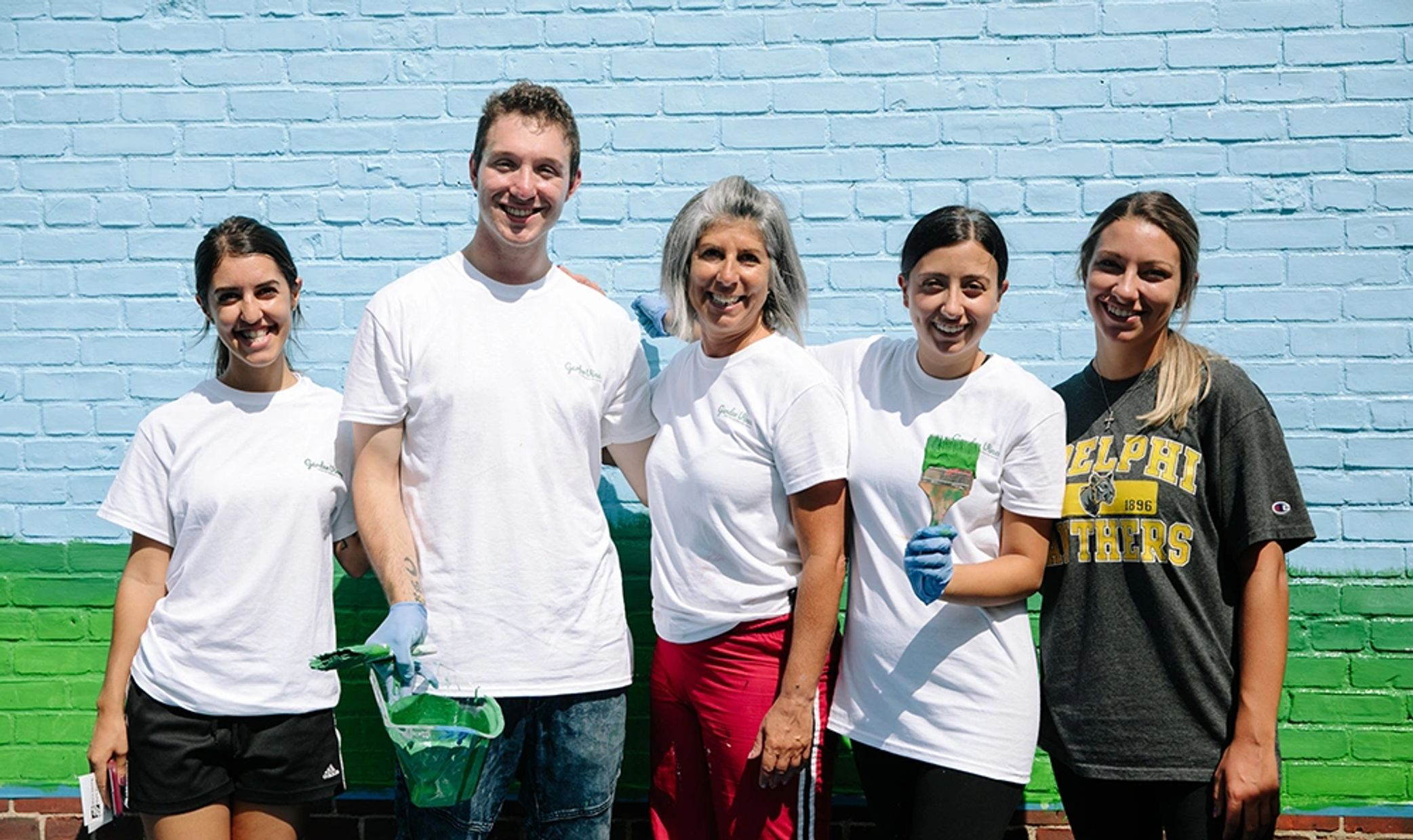 Group of five smiling people painting a brick wall with blue and green colors, holding paint brushes and a paint bucket, outdoors.