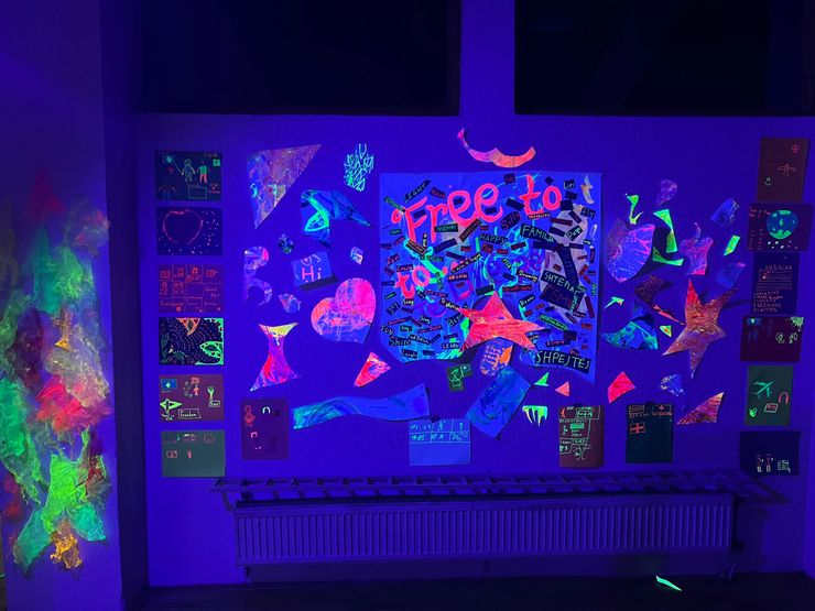 Colorful neon and glow-in-the-dark artwork on a classroom wall, with various shapes, drawings, and words illuminated under black light.