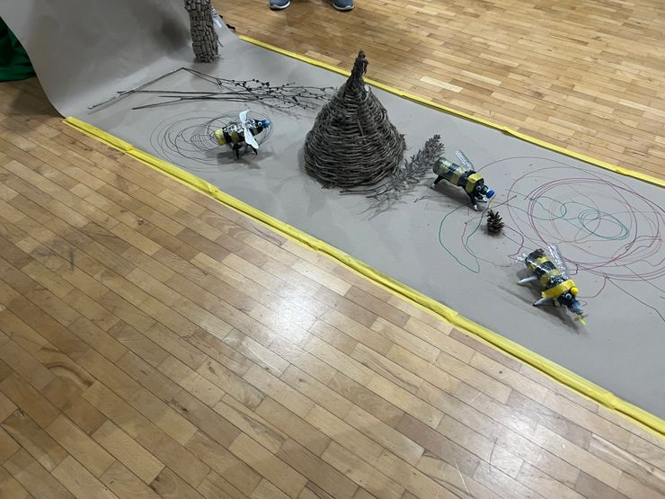 Art project display featuring a twisted wire cone, small pinecones, and four robots made from plastic bottles and toy parts, decorated with yellow and black tape, on a gray surface with drawn colorful spirals.