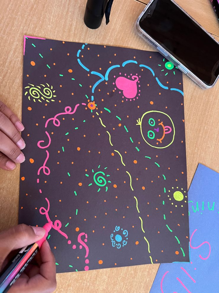 Neon-colored doodles on black paper, including a pink heart, a smiling face, swirls, a sun, and various lines and dots, with a person drawing with a pink marker.