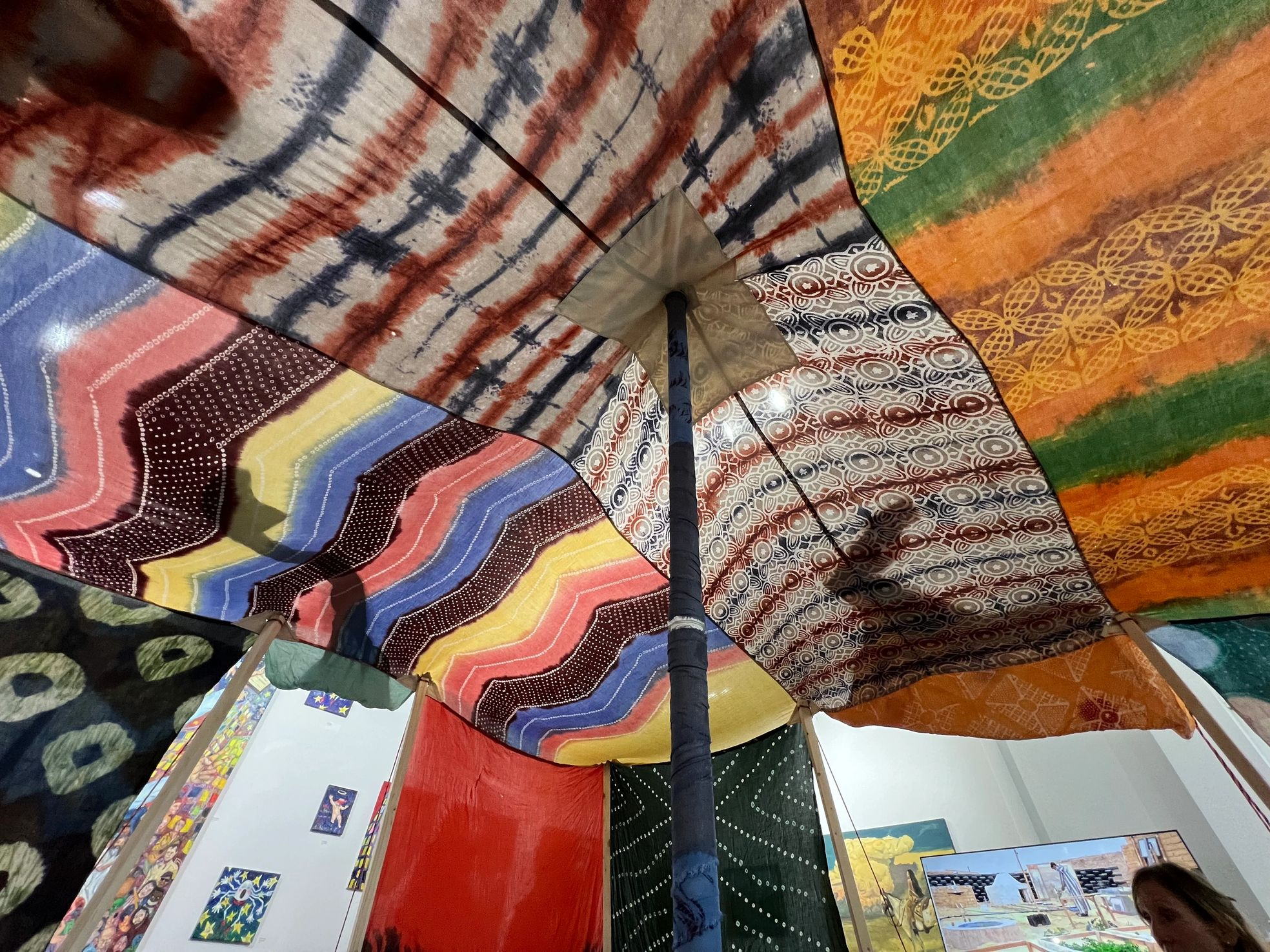 Colorful umbrella with various patterned sections, displayed indoors with artwork on the walls and a person partially visible in the foreground.