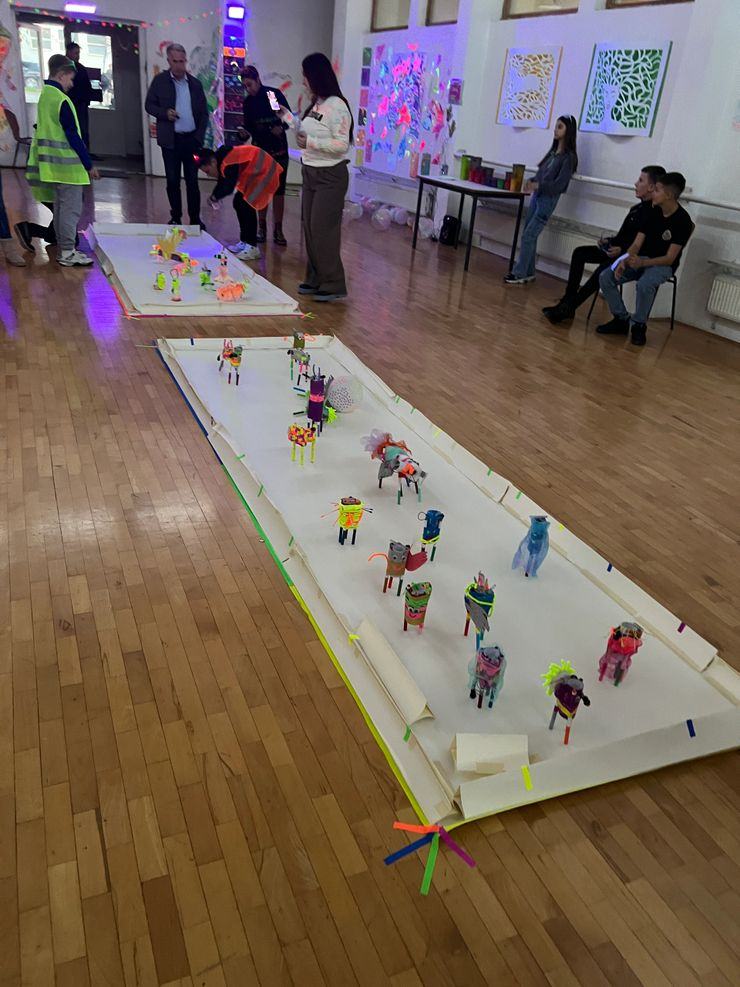 Room decorated with colorful artwork and paper animal crafts arranged on white sheets of paper on the floor, with people observing and taking photos.