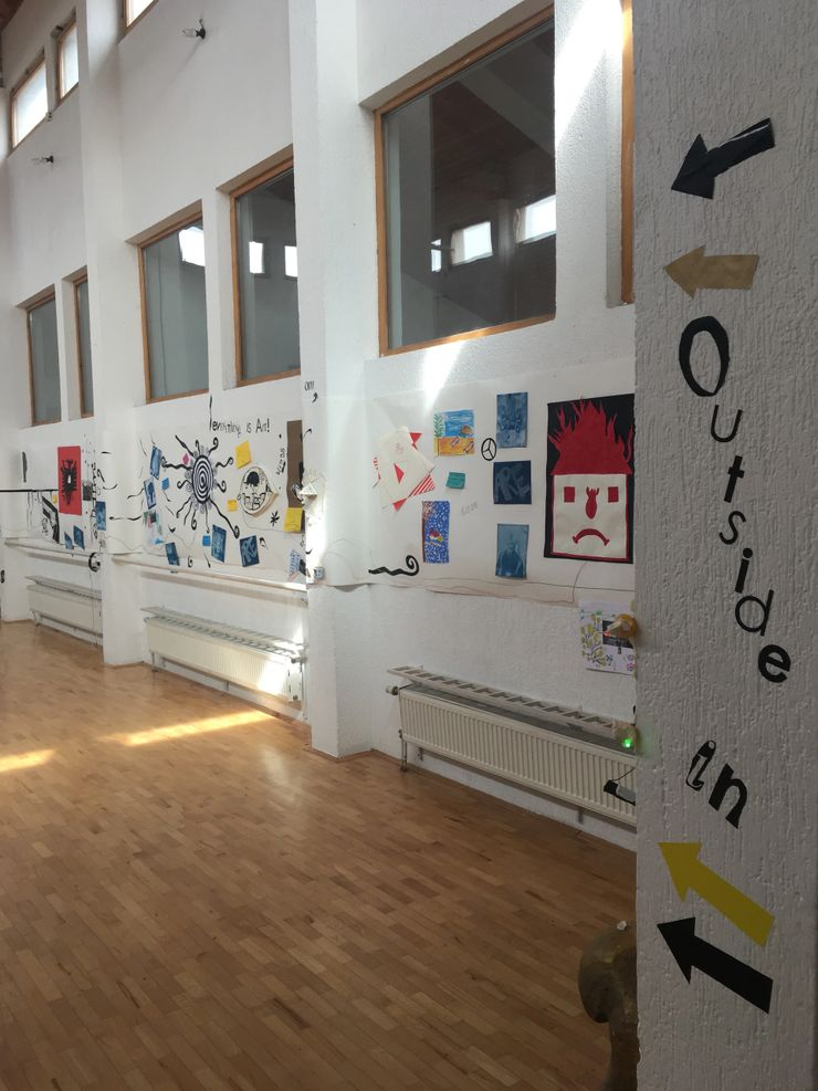 A hallway with wooden floors and white walls decorated with colorful artwork and drawings. There is an arrow sign on the wall pointing to the left with the text 'Outside in,' guiding visitors along the corridor.
