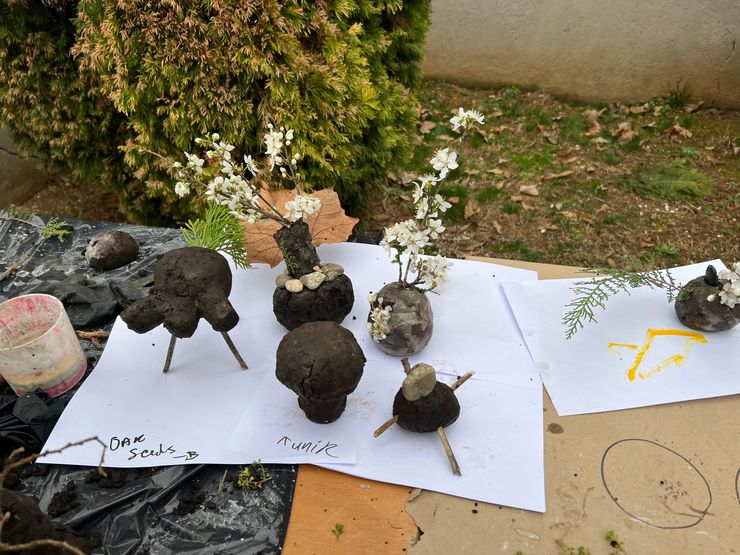 Small DIY plant sculptures made from rocks and soil, with white blossoms, on paper and cardboard outdoors.