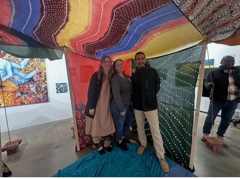 Three people are standing inside a colorful, patchwork-style tent made of fabric sheets, in an art gallery. The tent has a multicolored roof and decorated walls, with one person visible outside the tent holding a rope.