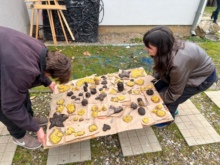 Two people are crouching down and looking at a large table outdoors, which is covered with various painted rocks in different shapes and sizes.