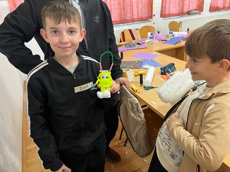 A young boy in a black Adidas jacket holding a green yarn craft with Christmas decorations, while another boy in a beige jacket looks on in a classroom with tables, chairs, and craft supplies.