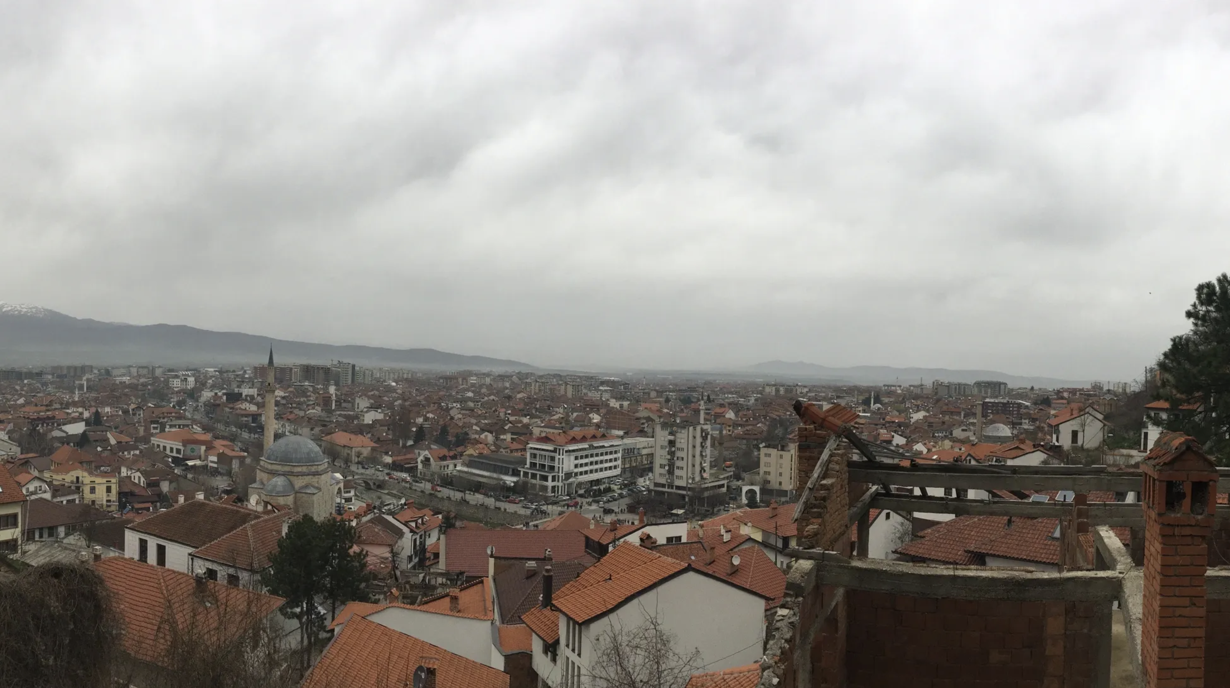 A panoramic cityscape view of a European city with red-tiled roofs, a mosque with minarets, modern buildings, and distant mountains under cloudy skies.
