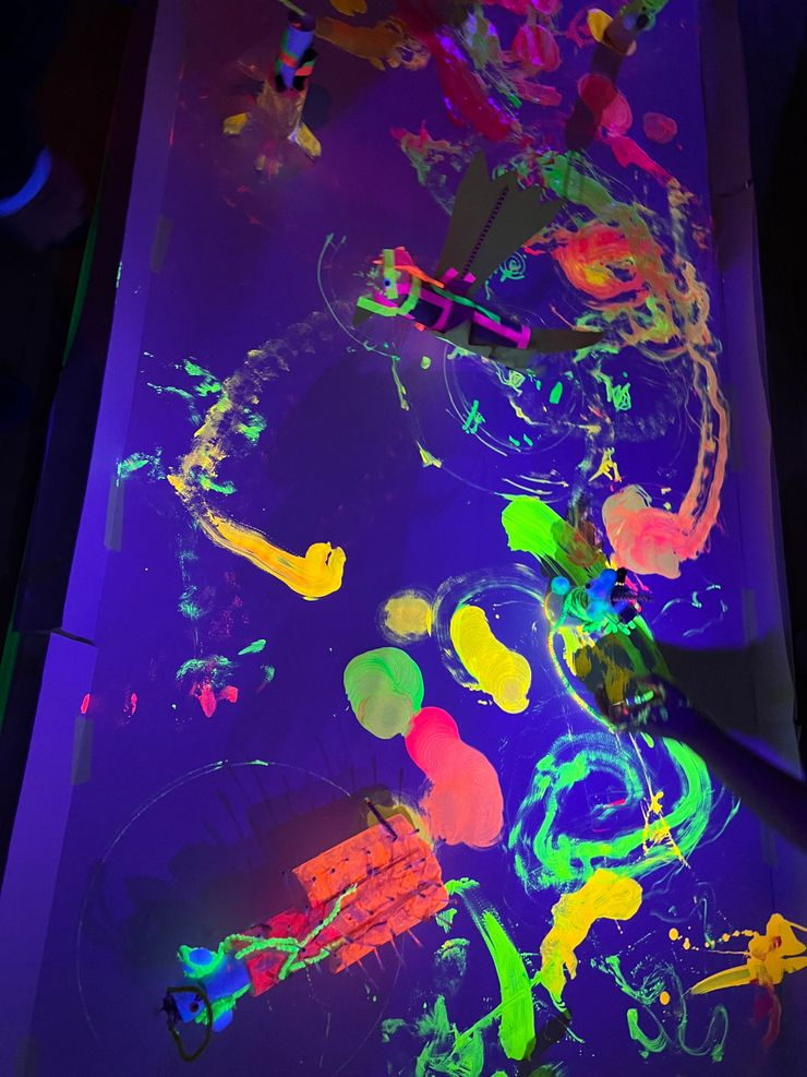 Colorful neon glow-in-the-dark painting with abstract shapes and figures on a dark surface.