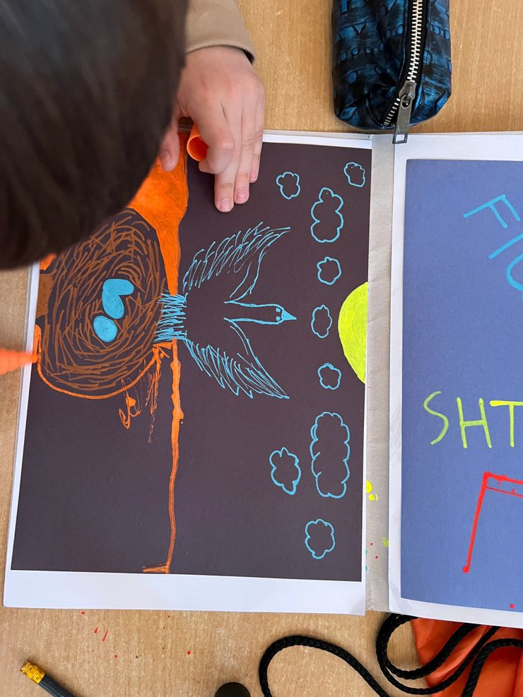Child drawing a scene of a wolf howling at the moon on black paper with blue, brown, and yellow chalk.