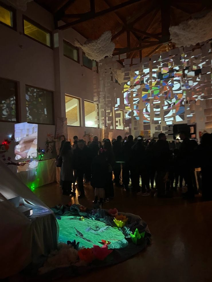 Indoor event with a crowd of people gathered in a dimly lit space, decorated with hanging paper or fabric art, and colorful projected patterns on a large wall, with some illuminated decorative elements on the floor.