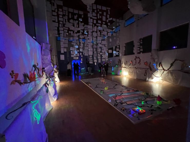 Dark room with glowing and decorated holiday or Christmas-themed decorations, including lights, paper crafts, and a large decorated display on the floor, with several people in the background.