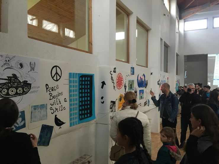 People observing artwork and posters on a gallery wall, including a peace sign, building graphic, tank drawing, and peace-related messages, in an indoor space with high ceilings and windows.