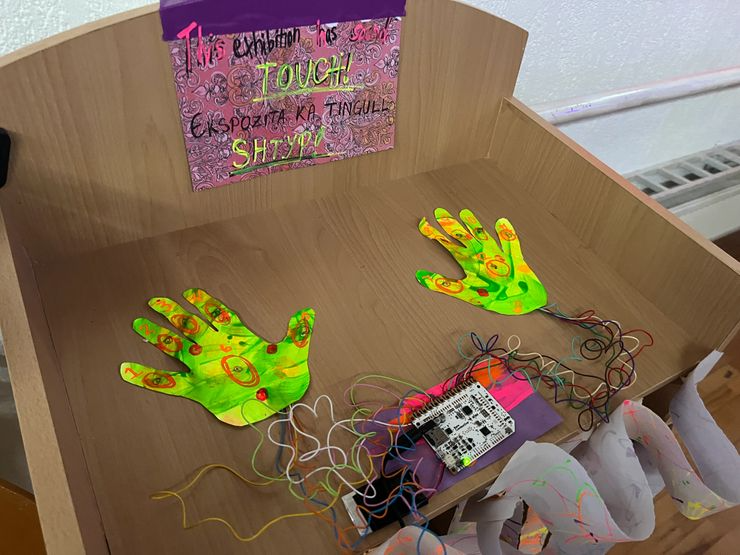 Interactive art display featuring two colorful, painted paper hands with wiring connected to electronic components on a wooden table, with a handwritten sign above that says 'This exhibition is to touch! Explore, compare, feel, and share!'