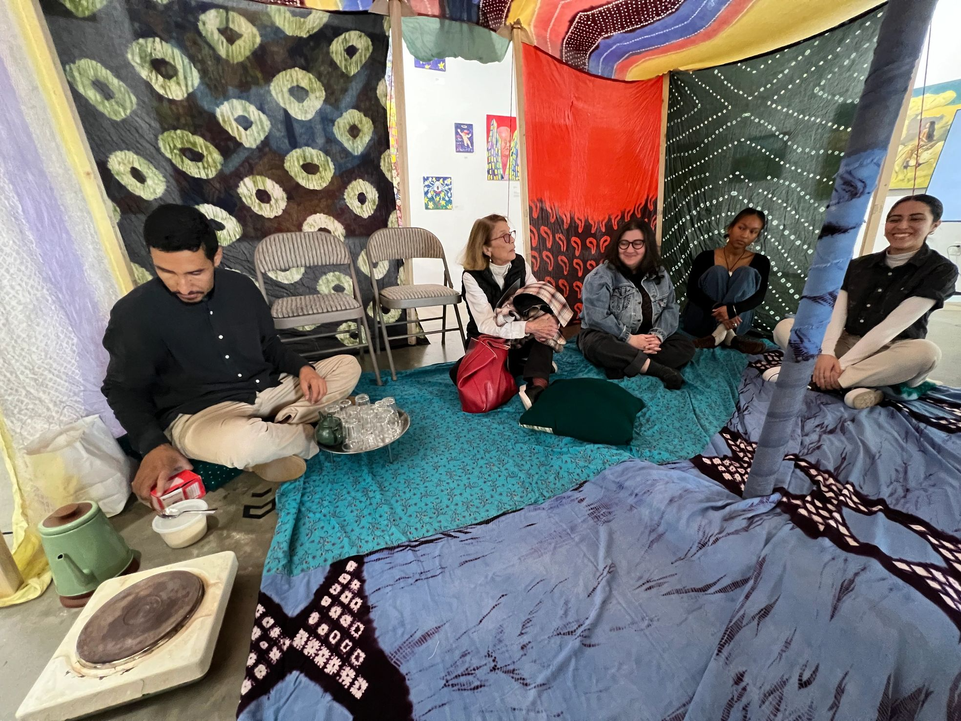 Group of five women sitting on colorful fabric-covered floor under vibrant, patterned fabric drapes in an art space with artworks on the walls. One woman is cooking or preparing food on a portable stove, and a tray of glasses and bottles are nearby.