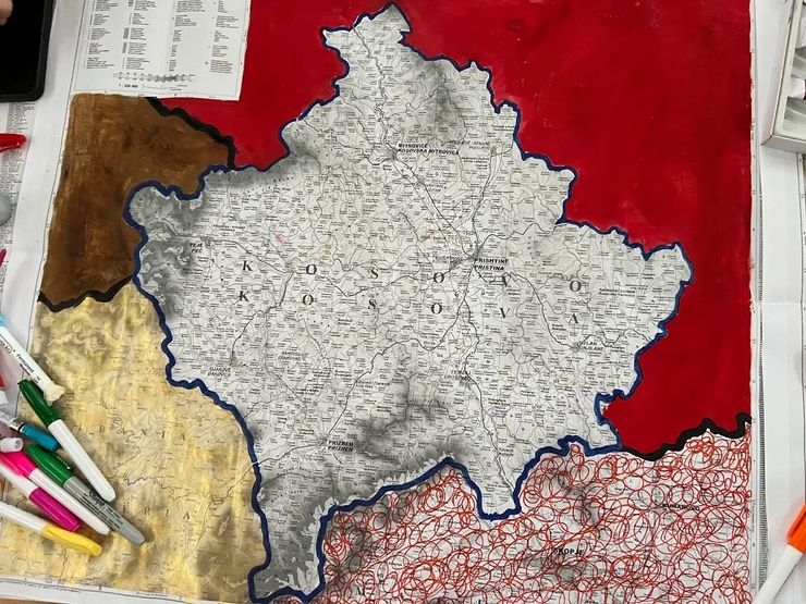 A paper map of Kosovo, outlined in blue, placed on a red background with various colored markers nearby.