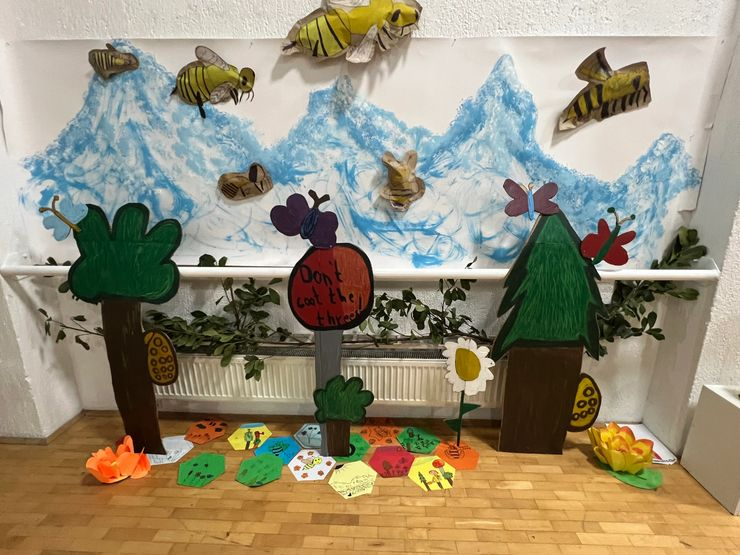 A children's craft display featuring paper trees, bee and butterfly cutouts, painted mountains in the background, and colorful paper cutouts on the floor with a sign that says, 'Don't eat the trees!'