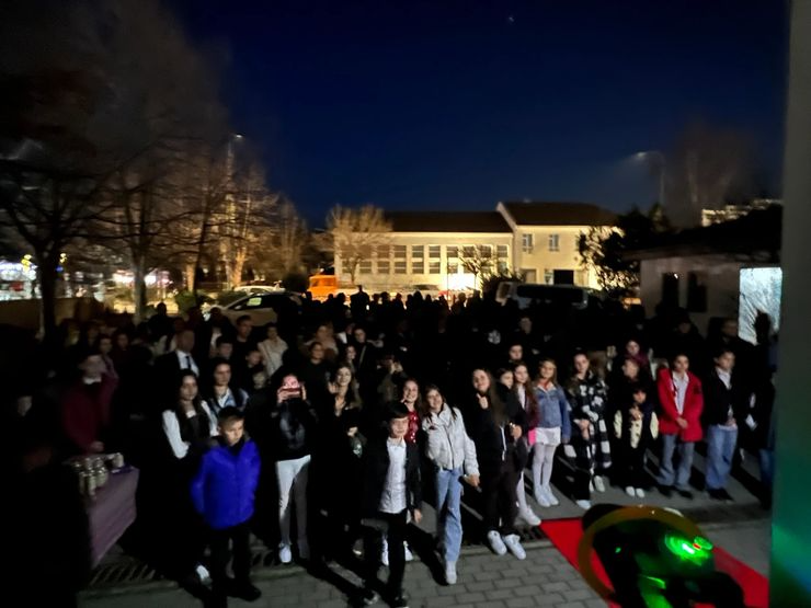 A large group of people, including children and adults, gathered outside at night, facing the camera. In the background, there are buildings, trees, and parked cars. The scene appears to be at an outdoor event or gathering.
