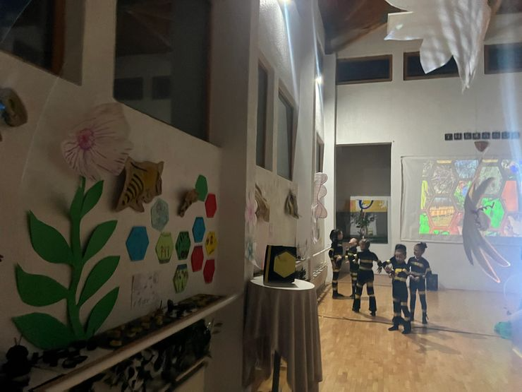 Classroom with children in striped uniforms holding hands, decorated with large paper flowers and artwork, sunlight streaming through windows.