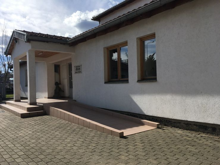 Exterior of a white building with a ramp and steps leading to the front door, three windows, and an overhang supported by two columns, with a paved walkway in front.
