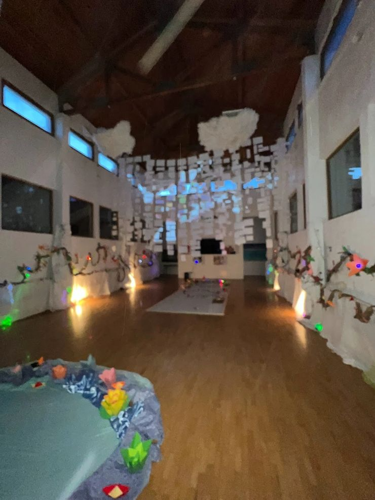 Indoor art installation with hanging paper artwork from ceiling, string lights, paper flowers, and clouds on the wall, creating a whimsical atmosphere.