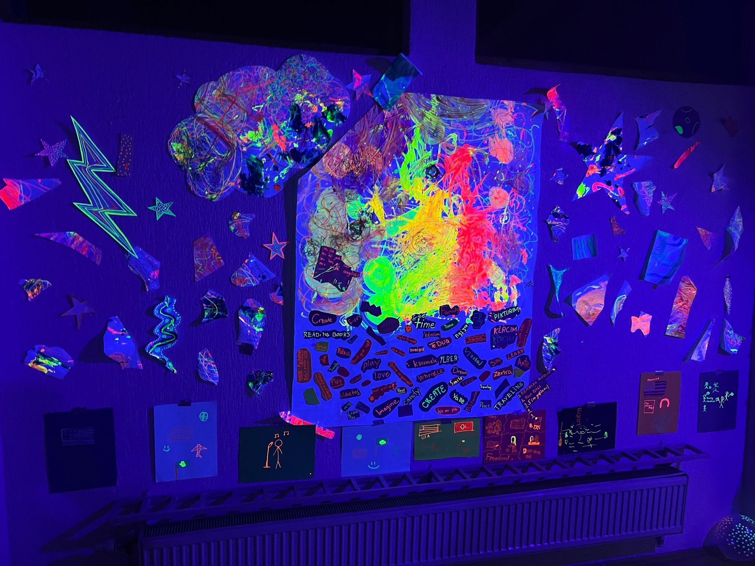Colorful artwork illuminated under black light, featuring a large abstract piece with bright neon colors, surrounded by paper cutouts of stars and shapes on a wall.