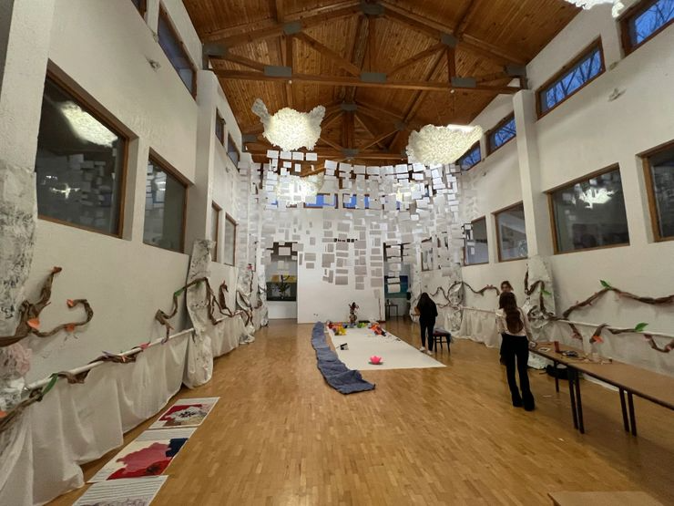 Indoor art exhibition with hanging paper structures, fabric-covered walls, and a long centerpiece on the floor with toys and decorations, in a wooden-ceiling gallery.