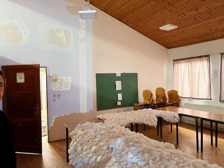 Classroom with a table covered in cotton, a green chalkboard on the wall, stacked chairs, windows with pink curtains, and a wooden ceiling.