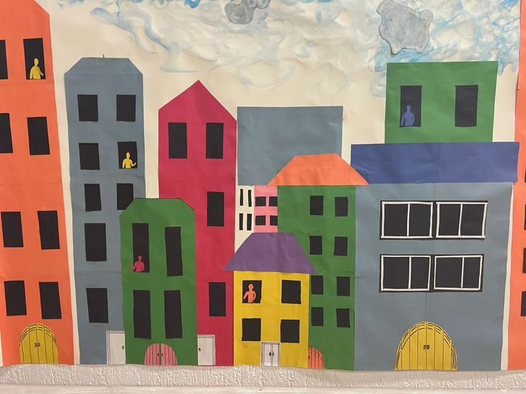 Colorful paper cutout cityscape with various buildings, windows, doors, and small painted people cutouts on the windows.