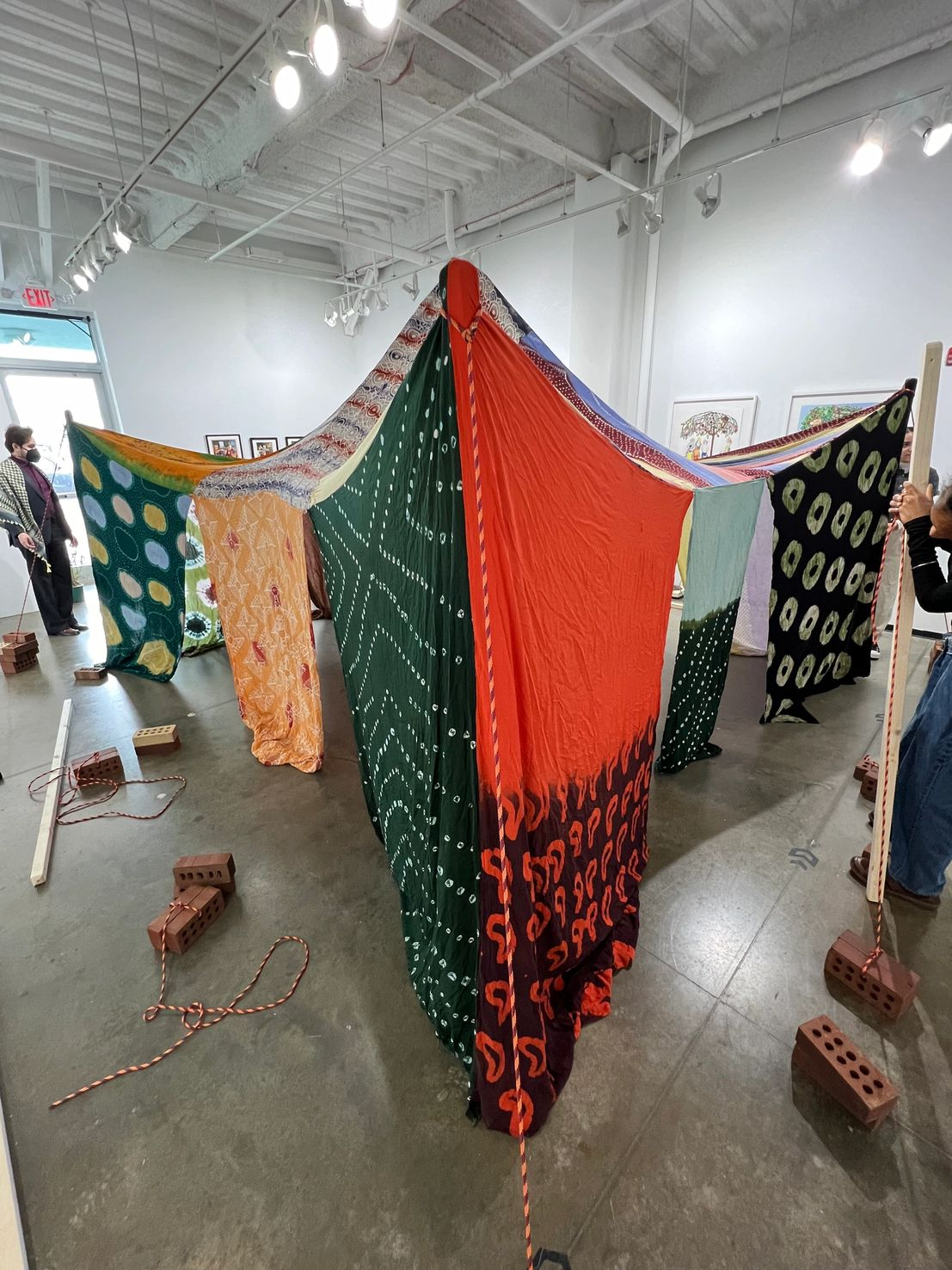An art installation in an gallery features a large, colorful fabric structure suspended from the ceiling. The fabric has various patterns and colors, including orange, green, purple, and black with designs. The structure is held down by ropes tied to