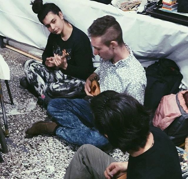 Three teenagers sitting on the floor in a casual setting, with one playing a small guitar or ukulele, others sitting nearby with backpacks and personal belongings.