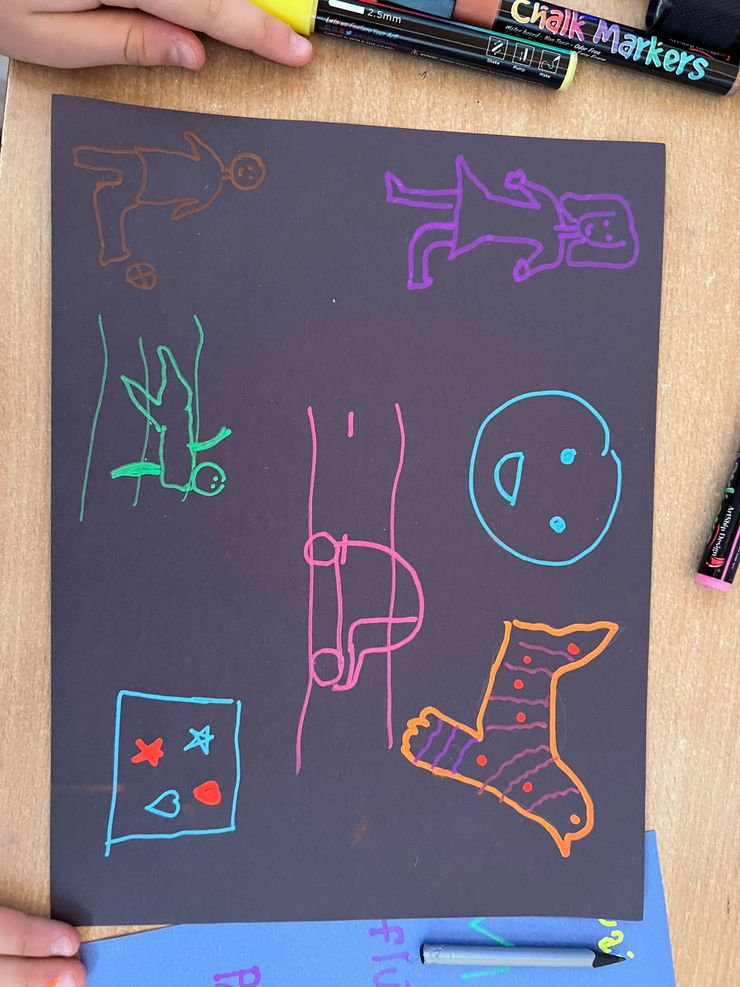 Colorful chalk drawings on black paper, including stick figures, a dog, a face, a sock, and a card with hearts and stars, surrounded by chalk markers.